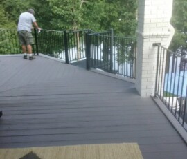 Person standing on a wooden deck next to a black metal railing, with trees and a white brick column in the background.