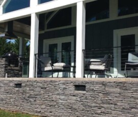 Modern house patio with stone retaining wall, white pillars, outdoor seating, and railing in front of large windows and doors.