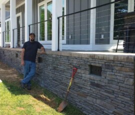 Man standing next to a stone retaining wall in front of a house with a porch, railing, and shovel in the ground nearby.