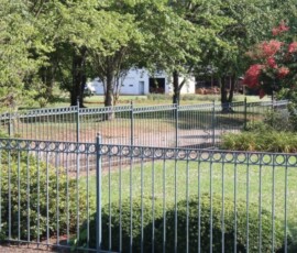 A black metal fence runs through a green lawn with bushes and trees, with a white building visible in the background.