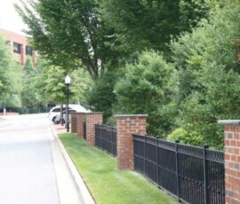 A brick and metal fence lines a sidewalk next to a street with trees, a white car, and a building in the background.