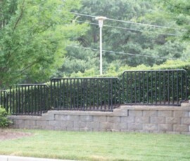 A black metal fence stands on a tiered stone wall with greenery and trees in the background, next to a parked car.