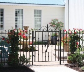 Black metal gate opens to a small courtyard with potted plants, a fountain, and white siding on the building.
