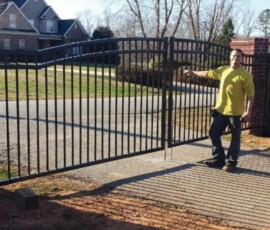 A person in a yellow shirt stands next to an open black metal gate at the entrance of a driveway.