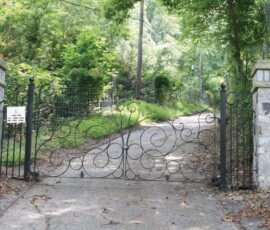 Ornate black iron gate with stone pillars blocks a paved driveway in a wooded, rural area.