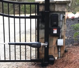 Black metal automatic gate with electronic opener attached to a stone post, set near a gravel driveway and trees.