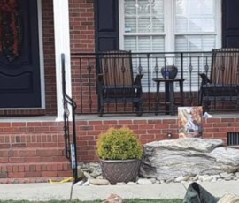 A brick porch with two chairs, a small table, potted plants, and a decorative rock in front of the house.