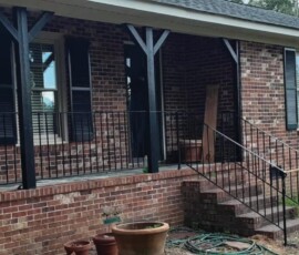 Brick house with a small porch, black railings, steps, and several empty flower pots and a garden hose in front.
